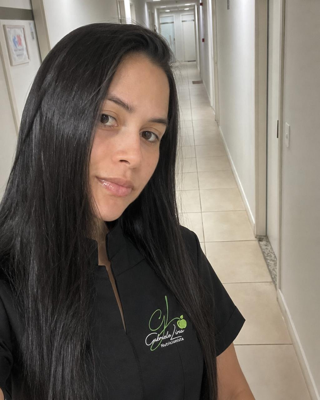 Gabriele Lima com uniforme profissional preto, com a sua marca, em corredor de ambiente clínico.